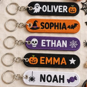 May include: Five rectangular embroidered keychains with metal rings. The black one features a ghost and pumpkin with the name OLIVER. The orange one has a witch hat and bat with SOPHIA. The purple one shows a skull and web with ETHAN. The black one has a pumpkin and star with EMMA. The white one displays a spider and candy with NOAH.