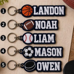 May include: Five embroidered fabric keychains with metal rings. Each features a sports ball icon and a name in white block letters on a dark blue background: a basketball for LANDON, football for NOAH, baseball for LIAM, soccer ball for MASON, and hockey puck for OWEN.