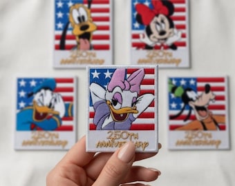 May include: A white tote bag with five embroidered patches featuring Disney characters. The patches showcase Pluto, Minnie Mouse, Donald Duck, Daisy Duck, and Goofy, each set against a red, white, and blue American flag design. The patches have the text "250th Anniversary".