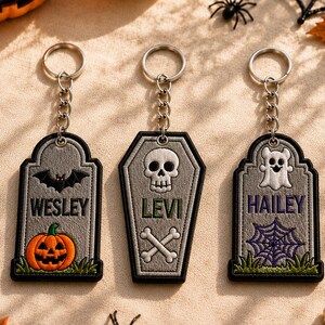 May include: Three embroidered keychains with metal rings. Two are gray tombstone shapes featuring a black bat with an orange pumpkin, and a white ghost with a purple spiderweb. The center keychain is a gray coffin shape with a white skull and crossbones.