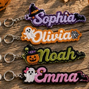 Puede incluir: Cuatro llaveros bordados con temática de Halloween y anillas de metal. El primero muestra el nombre Sophia en morado con un sombrero de bruja, el segundo Olivia en naranja con un fantasma, el tercero Noah en verde con una calabaza y el cuarto Emma en rosa con un fantasma.