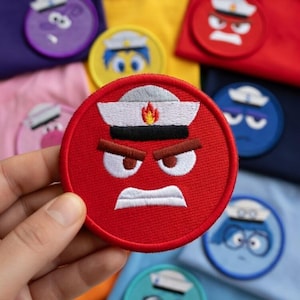 May include: A collection of embroidered patches depicting cartoon faces with various emotions. The patches are circular and vibrant, showcasing designs such as a red angry face, a yellow happy face, and a blue sad face. The patches are set against colourful fabric.