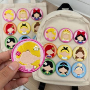 May include: Embroidered patches featuring cartoon princess designs. The patches are round with colorful details, including blonde, red, and black hair. Some patches are on a beige tote bag and backpack. One patch is held in a hand.