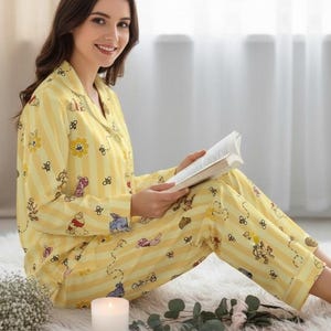 May include: Yellow pajama set with a Winnie the Pooh and flower pattern. The long-sleeved top and pants have vertical stripes. The person in the image is reading a book while sitting on a white rug.