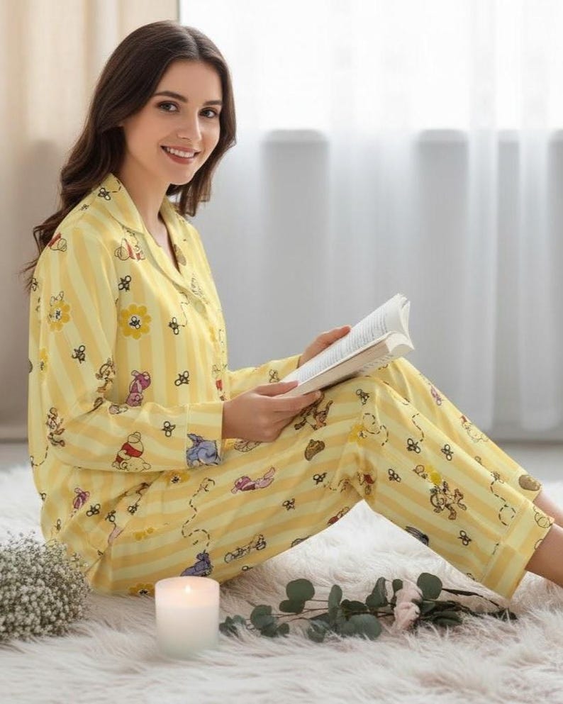 May include: Yellow pajama set with a Winnie the Pooh and flower pattern. The long-sleeved top and pants have vertical stripes. The person in the image is reading a book while sitting on a white rug.