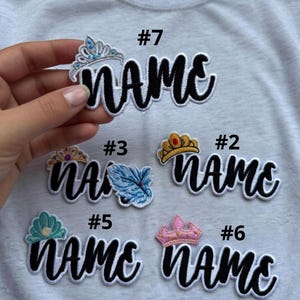 May include: Embroidered patches with the word "NAME" in a black script font, each topped with a different crown design. The patches are in various colors, including gold, pink, and blue, and are displayed on a white t-shirt.
