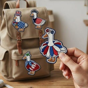 Könnte beinhalten: Ein beigefarbener Canvas-Rucksack mit braunen Lederriemen und drei gestickten Gänseaufnähern. Ein Aufnäher wird von einer Hand gehalten. Die Aufnäher zeigen Gänse in patriotischen Outfits mit roten, weißen und blauen Farben. Die Aufnäher sind dekorativ.
