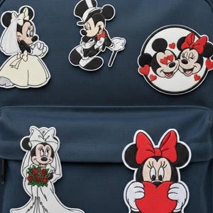 May include: A navy blue backpack adorned with embroidered patches of Mickey and Minnie Mouse. The patches show Mickey in a tuxedo, Minnie in a wedding dress, and Minnie holding a red heart.