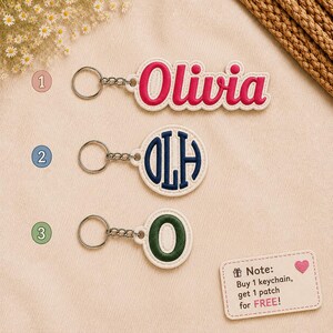 May include: Three custom keychains with metal rings. The first is a white patch with the pink embroidered name Olivia. The second is a circular white patch with a dark blue embroidered monogram OLH. The third is a circular white patch with a dark green embroidered letter O. A small rectangular sign includes the text Note: Buy 1 keychain, get 1 patch for FREE.