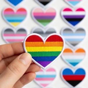 May include: A collection of heart-shaped patches with various LGBTQ+ pride flag designs. One patch, held by a hand, displays a rainbow flag. The patches are embroidered with a white border and are set against a white background.