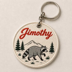 以下が含まれることがあります： 赤い筆記体でJimothyという名前が刺繍された円形のキーホルダー。背景には緑の松の木と山の頂上が描かれ、その前を歩く灰色のタヌキがデザインされており、銀色の金属製リングに取り付けられています。