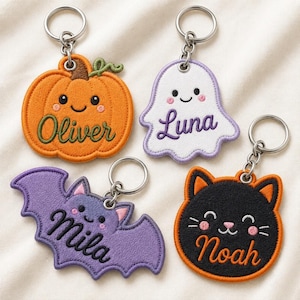 Op de afbeelding: Vier geborduurde sleutelhangers in Halloween-vormen: een oranje pompoen met de naam Oliver, een wit spookje met de naam Luna, een paarse vleermuis met de naam Mila en een zwarte kat met een oranje rand en de naam Noah.