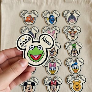 May include: A collection of character-themed stickers in the shape of Mickey Mouse ears. The stickers feature various cartoon characters, the year "2020" or "2026", and the word "Name". The stickers are on a beige tote bag.