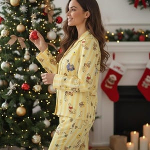 May include: Yellow pajama set with a Winnie the Pooh pattern. The set includes a button-down top and matching pants with vertical stripes. The background features a decorated Christmas tree and a fireplace with stockings.