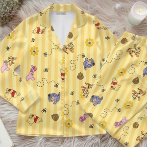 May include: Yellow pajama set with vertical stripes and cartoon character pattern. The long-sleeved top has a collared neckline and button closure. The matching pants are visible. The set features Winnie the Pooh characters, flowers, and honey pots.