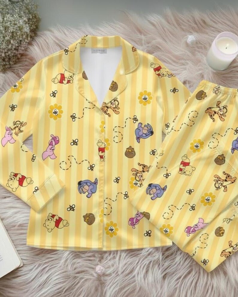 May include: Yellow pajama set with vertical stripes and cartoon character pattern. The long-sleeved top has a collared neckline and button closure. The matching pants are visible. The set features Winnie the Pooh characters, flowers, and honey pots.