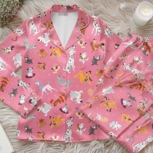 May include: Pink pajama set with a repeating pattern of various dog breeds. The long-sleeved top has a notched collar and white trim. The matching pants are folded. The set is on a fluffy, white surface.