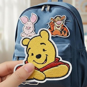 Può includere: Uno zaino blu decorato con toppe ricamate di Winnie the Pooh, Tigro e Pimpi. Una toppa di Winnie the Pooh è tenuta in mano, mostrando il volto sorridente dell'orso giallo e la sua camicia rossa. Lo zaino ha una tasca frontale e una maniglia superiore.