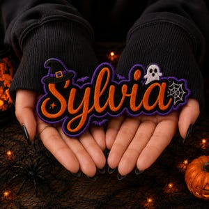 May include: An embroidered patch featuring the name Sylvia in orange cursive script with a thick purple border. The design includes a black witch hat, a small white ghost, a spider web, and tiny purple bats, all set against a dark purple background.