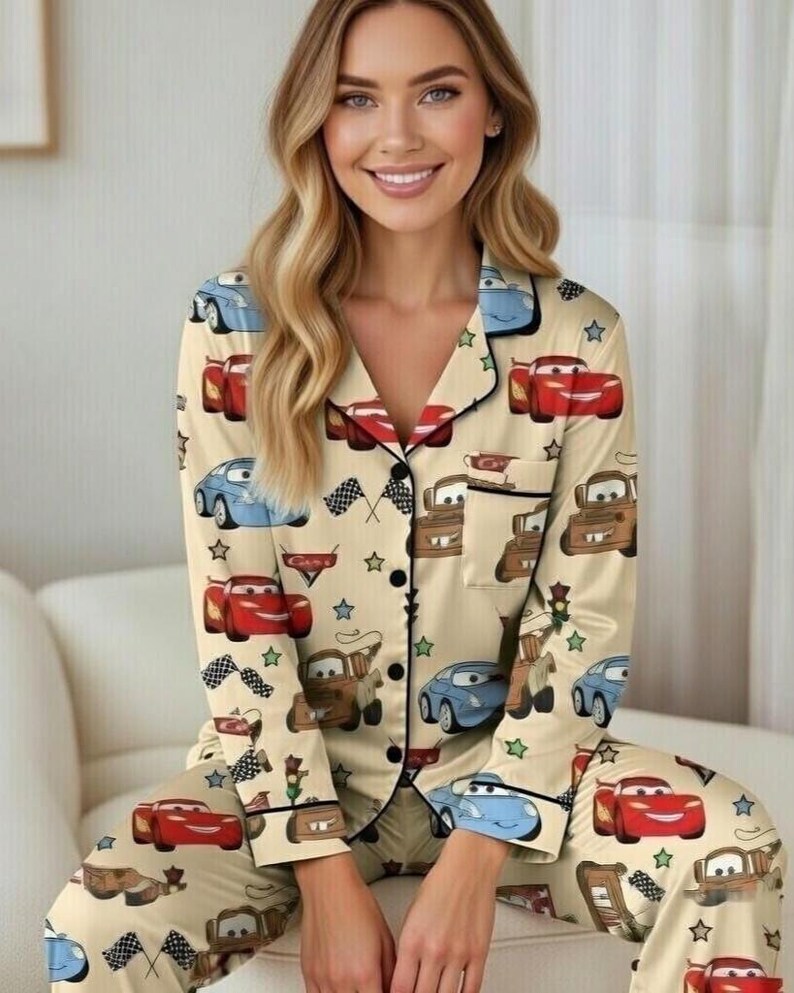 Op de afbeelding: Beige pyjamaset met een herhalend patroon van cartoonauto's, vlaggen en sterren. De top met lange mouwen heeft een reverskraag en zwarte bies. De bijpassende broek maakt de set compleet. De pyjama is gemaakt van zacht materiaal.