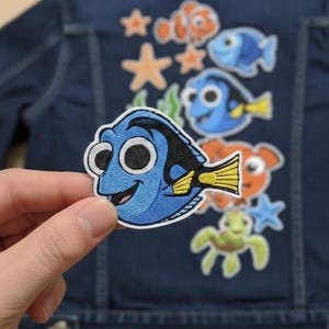 Puede incluir: Un parche bordado azul de Dory de la película Buscando a Nemo, sostenido frente a una chaqueta de mezclilla con otros parches bordados de criaturas marinas. El parche de Dory es azul con cola amarilla y borde blanco.