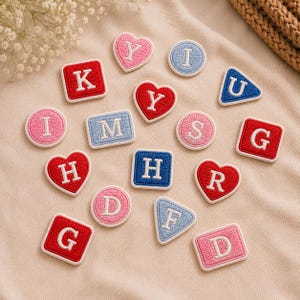 Op de afbeelding: Een verzameling geborduurde stoffen patches in verschillende vormen, waaronder harten, cirkels, vierkanten en driehoeken. Elke patch heeft een witte letter in het midden, met kleuren zoals rood, lichtblauw en roze met witte randen.