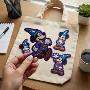 Puede incluir: Parches bordados con Mickey Mouse con un disfraz de mago. Los parches están en varias poses y colores, incluyendo morado, azul y marrón. Los parches están sobre una bolsa de mano beige.