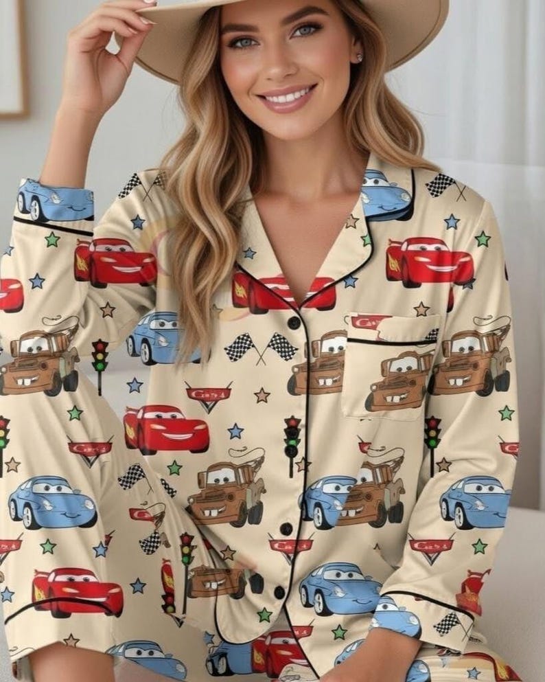 Op de afbeelding: Beige pyjamaset met een herhaald patroon van cartoonauto's, verkeerslichten en geblokte vlaggen. De top met lange mouwen heeft zwarte biezen en een borstzak. De pyjamaset is gemaakt van een zacht, comfortabel materiaal.