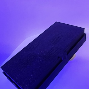 Könnte beinhalten: Ein rechteckiges, schwarzes Objekt mit strukturierter Oberfläche und erhabenem Rand. Das Objekt ist abgewinkelt, mit einem kleinen, abgerundeten Detail auf einer Seite. Der Hintergrund ist ein Blau-Weiß-Verlauf.