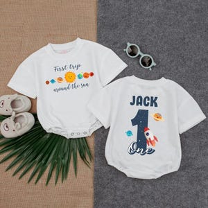 May include: Two white baby onesies. One has the text "First trip around the sun" with a sun and planet graphic. The other has the name "Jack", the number "1", a rocket, and the word "One".