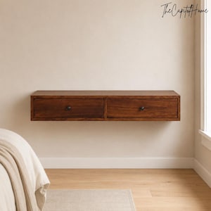May include: A wall-mounted floating shelf made of dark brown wood featuring two horizontal drawers with small, round, dark-coloured knobs. The unit has a rectangular shape and a smooth finish, positioned horizontally against a plain wall.