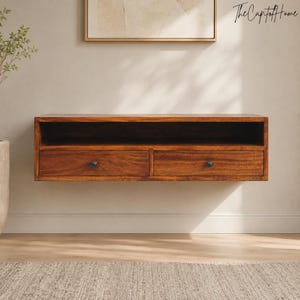 May include: A rectangular wall-mounted wooden shelf featuring a dark brown wood grain finish. The unit includes an open top storage compartment and two lower drawers, each equipped with a single small dark circular knob for opening and closing.