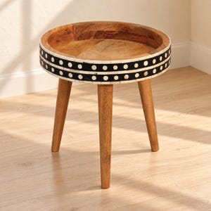 May include: A round wooden side table with three tapered legs. The circular tabletop features a raised edge and a decorative band around the side, showcasing a black and white pattern of alternating dots and horizontal lines.