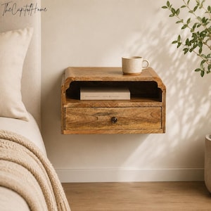 Peut inclure: Une table de chevet flottante en bois fixée au mur avec une finition à grain naturel, comprenant une étagère de rangement ouverte contenant un livre et un tiroir unique avec un petit bouton sombre. Une tasse en céramique beige est posée dessus.