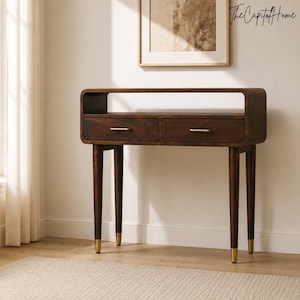 Puede incluir: Una mesa consola de madera marrón oscuro con un marco rectangular curvado, un estante superior abierto y dos cajones con tiradores horizontales de metal dorado. La mesa se apoya sobre cuatro patas cónicas, cada una con una base metálica dorada.