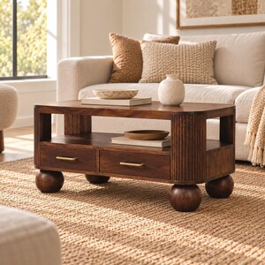 Peut inclure: Une table basse rectangulaire en bois foncé dotée de deux tiroirs avec de fines poignées horizontales dorées, d'une étagère inférieure ouverte et de quatre grands pieds sphériques. La table présente des coins arrondis et des cannelures verticales.