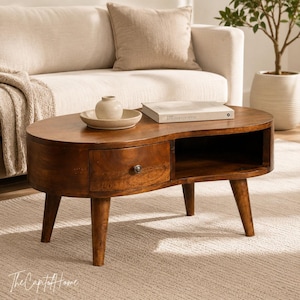 May include: A kidney-shaped wooden coffee table with a rich brown finish, featuring one central drawer with a small round knob and an open storage compartment. The table is supported by four tapered legs and holds a small ceramic bowl and a book.