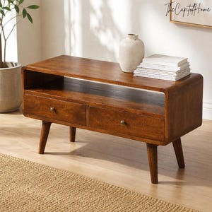 Peut inclure: Un meuble télé rectangulaire en bois au fini brun chaud, doté de deux tiroirs avec de petits boutons ronds et d'une étagère de rangement ouverte au-dessus. L'unité repose sur quatre pieds fuselés et présente des bords arrondis sur les côtés.