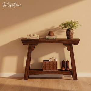 Op de afbeelding: Een donkerbruine houten console met een onderste plank. De bovenste plank bevat een beige gevouwen item, een kleine vaas, boeken en een decoratieve vaas met groen. Een gevlochten mand en bruine laarzen staan op de onderste plank. De tekst "The Capitol Home" staat linksboven.