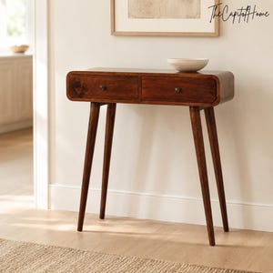 Op de afbeelding: Een donkerbruine houten sidetable met twee lades, afgeronde hoeken en vier taps toelopende poten. Een kleine, lichtgekleurde kom staat op het oppervlak. De tafel heeft een gladde afwerking en eenvoudige metalen knoppen op de ladefronten.