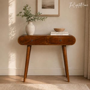 May include: A mid-century modern wooden console table with two drawers and tapered legs. A white vase with greenery, a wooden bowl, and books are displayed on the table. The Capital Home logo is in the upper right corner.