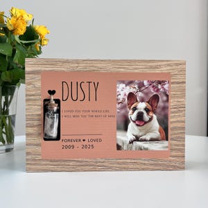 Può includere: Una cornice rettangolare in legno con finitura a venature color marrone chiaro. La cornice contiene una foto di un bulldog francese e una targa commemorativa con il testo DUSTY, I LOVED YOU YOUR WHOLE LIFE, I WILL MISS YOU THE REST OF MINE, FOREVER LOVED e 2009-2025. Una piccola fiala di vetro con tappo di sughero è fissata sul lato sinistro.