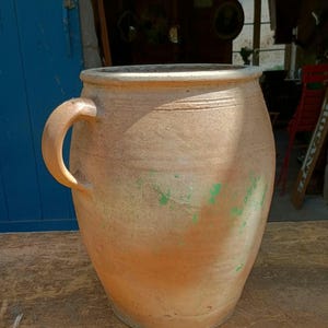 Pode incluir: Um grande pote de terracota antigo com uma alça. O pote tem uma boca larga e um corpo arredondado. É de cor castanho claro com manchas de tinta verde. O pote é provavelmente usado para armazenamento ou decoração.