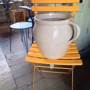 以下が含まれることがあります： 明るい黄色の折りたたみ椅子の上に、シングルハンドルが付いた大きなオフホワイトのセラミックジャグが置かれています。椅子は木と金属でできています。背景には、青い金属製の椅子、テーブル、木の床が含まれています。