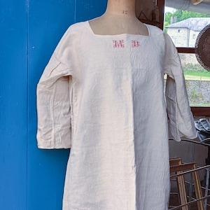 Peut inclure: Robe en lin crème avec un col carré et des manches trois-quarts. La robe présente des initiales brodées en rouge sur la poitrine. Le vêtement est présenté sur un mannequin sur fond bleu.