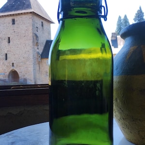 Op de afbeelding: Een hoge, cilindrische groene glazen fles met een draadbeugelsluiting aan de bovenkant. De fles heeft een glad, doorschijnend oppervlak en een afgeronde hals, ontworpen voor opslag of decoratief gebruik, rechtopstaand met een helder glazen lichaam.
