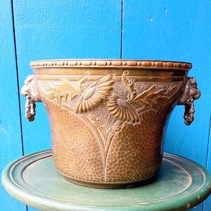 Può includere: Un vaso di fiori color bronzo con una superficie strutturata e rilievi floreali. Il vaso presenta due manici a forma di testa di leone e un bordo decorativo. Il design include girasoli e foglie. Il vaso è appoggiato su un tavolo verde.