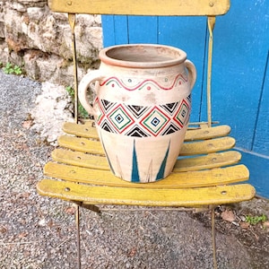 May include: A cream-coloured ceramic vase with two handles, decorated with geometric patterns in red, black, and green. The vase sits on a yellow folding chair against a blue door and stone wall.