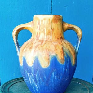 May include: A ceramic vase with two handles, featuring a color scheme of blue, yellow, and brown. The vase has a bulbous shape with a blue base, transitioning to yellow and brown hues towards the top. The handles are curved.