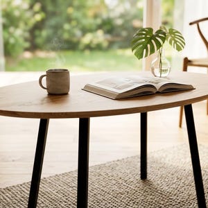 Peut inclure: Une table basse ovale marron clair avec des pieds noirs. Une tasse fumante, un livre ouvert et un vase avec des feuilles vertes sont posés sur la table. Une chaise en bois est à l'arrière-plan, et la table repose sur un tapis gris.
